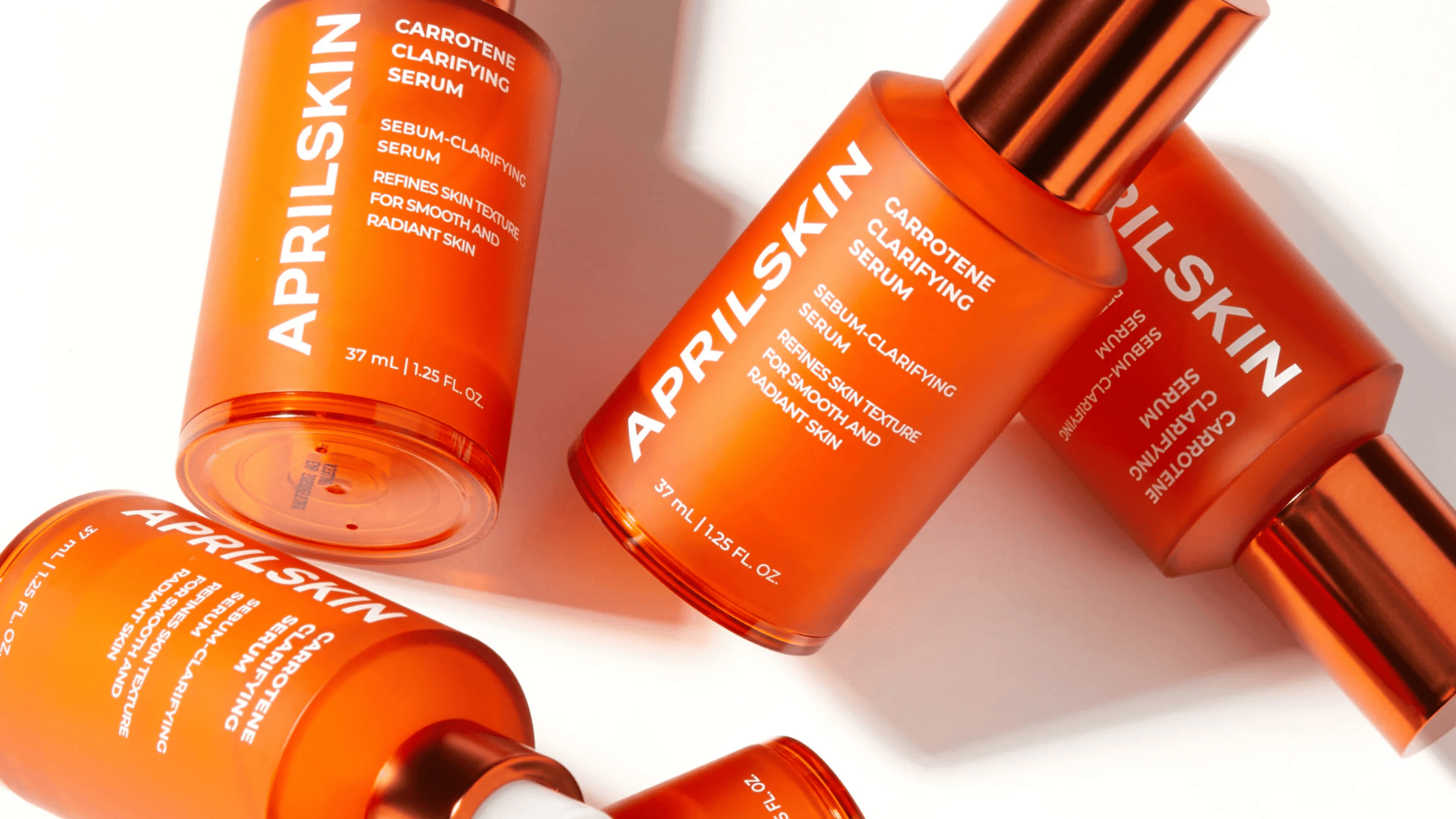 Bilden visar ett sortiment av APRILSKIN hudvårdsprodukter, specifikt deras "Carrotene Clarifying Serum." Flaskorna är enhetligt utformade med en iögonfallande orange färg, vilket speglar ingrediensen morot som är känd för sina hudklarande egenskaper.