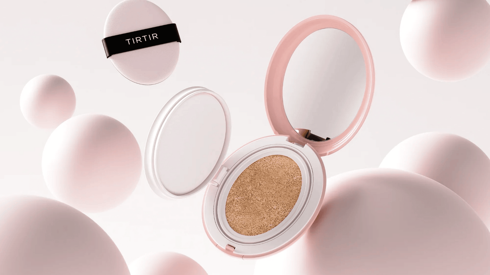 Bilden visar en öppen cushion foundation med märket "TIRTIR" på locket. Produkten är placerad mot en bakgrund av abstrakta vita och rosa sfärer som skapar en mjuk och ren atmosfär, vilket antyder en skönhetsprodukt med en elegant och modern design.