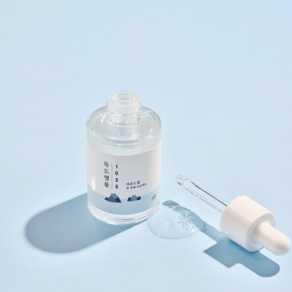 Una bottiglia aperta di 1025 Dokdo Ampoule con un contagocce accanto, su uno sfondo blu che crea un'ombra morbida.