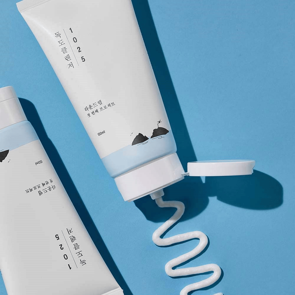 Due tubi di 1025 Dokdo Cleanser su sfondo blu, uno con tappo aperto e una striscia ondulata di detergente spremuta fuori.