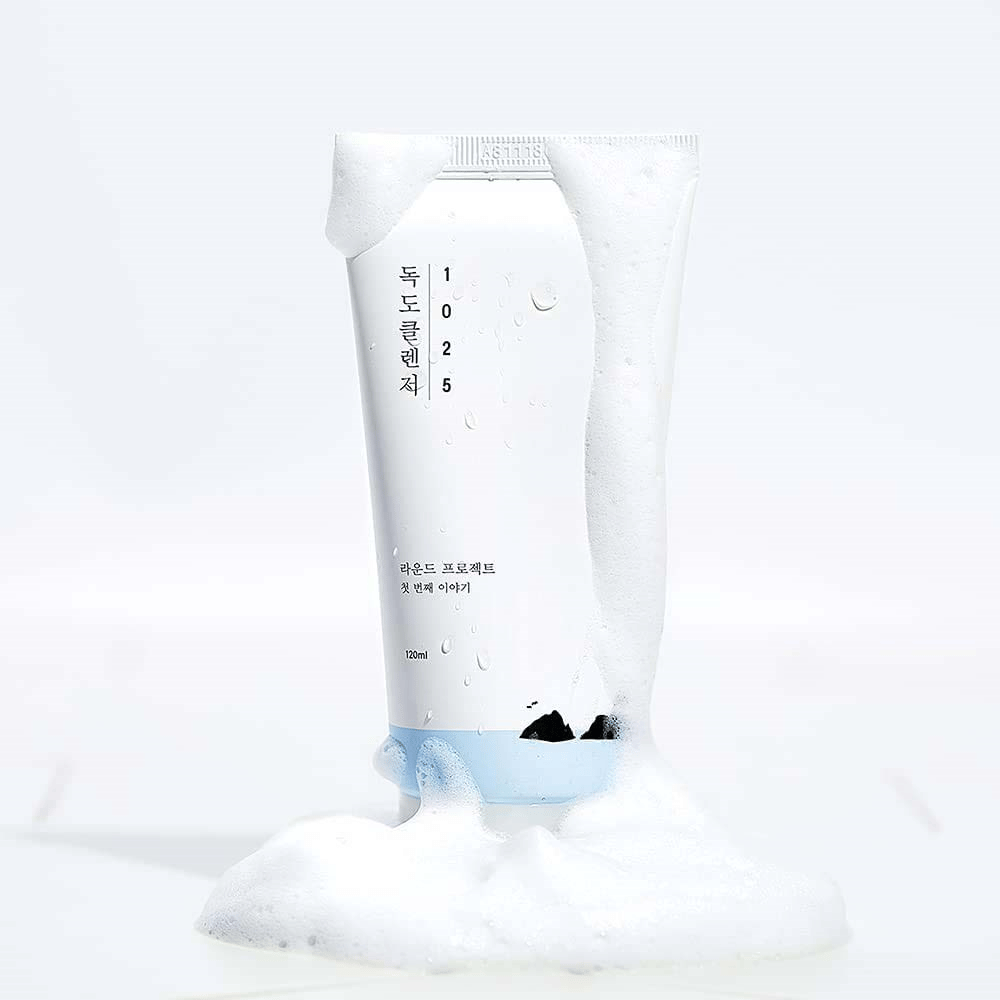 1025 Dokdo Cleanser i vit tub täckt av lödder, med vattenpärlor och skum runt botten, frilagd mot ljus bakgrund.
