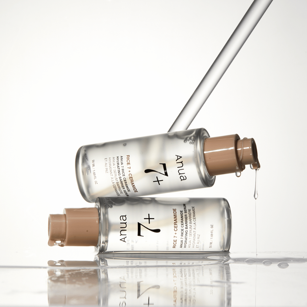 L'immagine mostra due bottiglie trasparenti di "Anua 77 Rice + Ceramide Hydrating Barrier Serum" con gocce che scorrono da una pipetta. Il siero, arricchito con riso e ceramidi, è destinato a idratare e rafforzare la barriera cutanea. Le bottiglie sono elegantemente progettate, il che rafforza la sensazione premium del prodotto.
