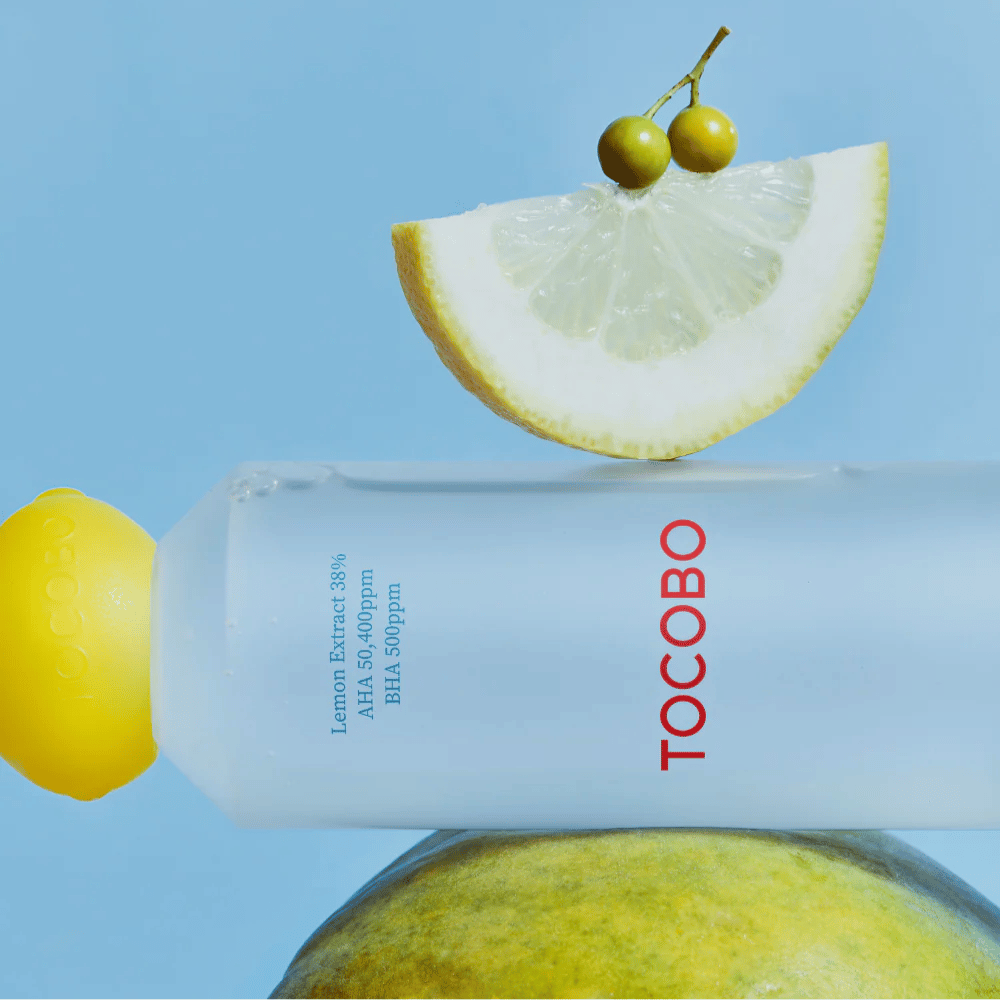 Bottiglia con TOCOBO AHA BHA Lemon Toner, decorata con un tappo a forma di limone. Una fetta di limone e piccole bacche verdi bilanciano sopra la bottiglia su uno sfondo blu.