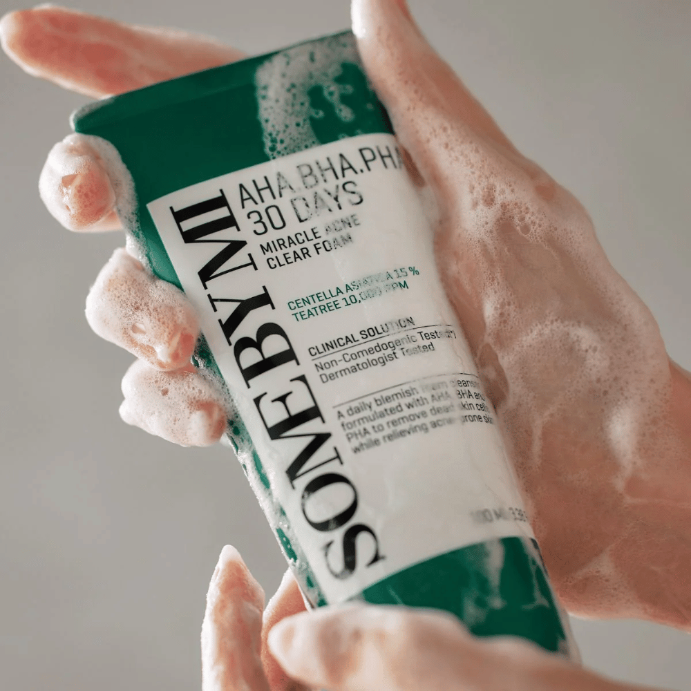 Una persona tiene in mano un tubo di "SOME BY MI AHA, BHA, PHA 30 Days Miracle Acne Clear Foam" con contenuto schiumogeno sopra le mani. Il prodotto è destinato all'uso quotidiano per combattere l'acne e pulire i pori con i suoi ingredienti attivi come centella, olio di tea tree e acidi come AHA, BHA e PHA.