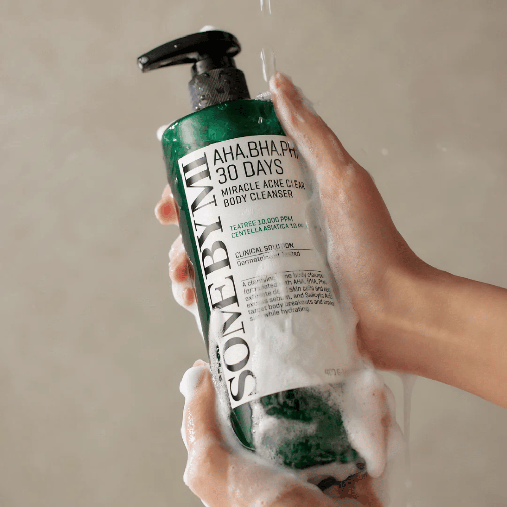 Una persona tiene in mano una bottiglia SOME BY MI AHA.BHA.PHA 30 Days Miracle Acne Clear Body Cleanser. La bottiglia è verde con un'etichetta bianca ed è coperta di schiuma dal detergente. La persona si lava con il prodotto mentre l'acqua scorre sulle mani.