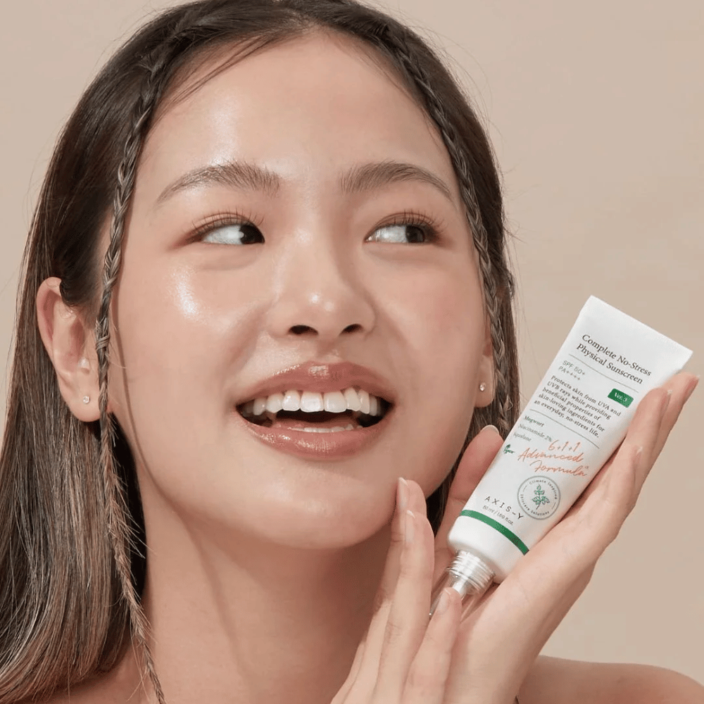 L'immagine mostra una giovane donna che sorride felicemente e tiene un tubetto di AXIS-Y Complete No-Stress Physical Sunscreen. Ha un aspetto radioso e naturale con un trucco minimo, che evidenzia il prodotto che presenta.