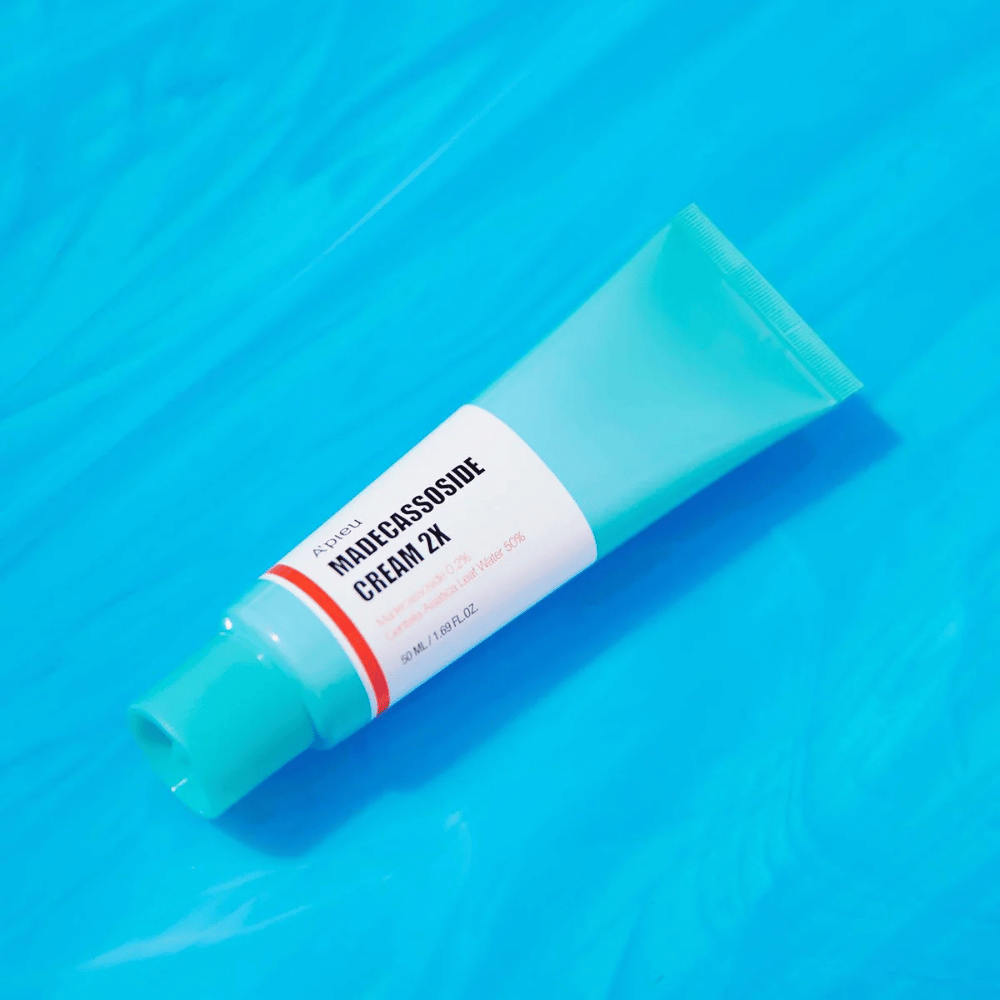 Un tubo A'PIEU Madecassoside Cream 2X è posizionato diagonalmente su uno sfondo blu con rughe simili all'acqua. Il tubo ha un corpo bianco con un tappo turchese chiaro e un'etichetta colorata dal rosso all'arancione dove il nome del prodotto e l'ingrediente attivo Madecassoside 2% sono chiaramente stampati. Sull'etichetta è indicata anche la quantità che è 50 ml / 1.8 fl oz. La presentazione luminosa e colorata dà una sensazione fresca ed energica.