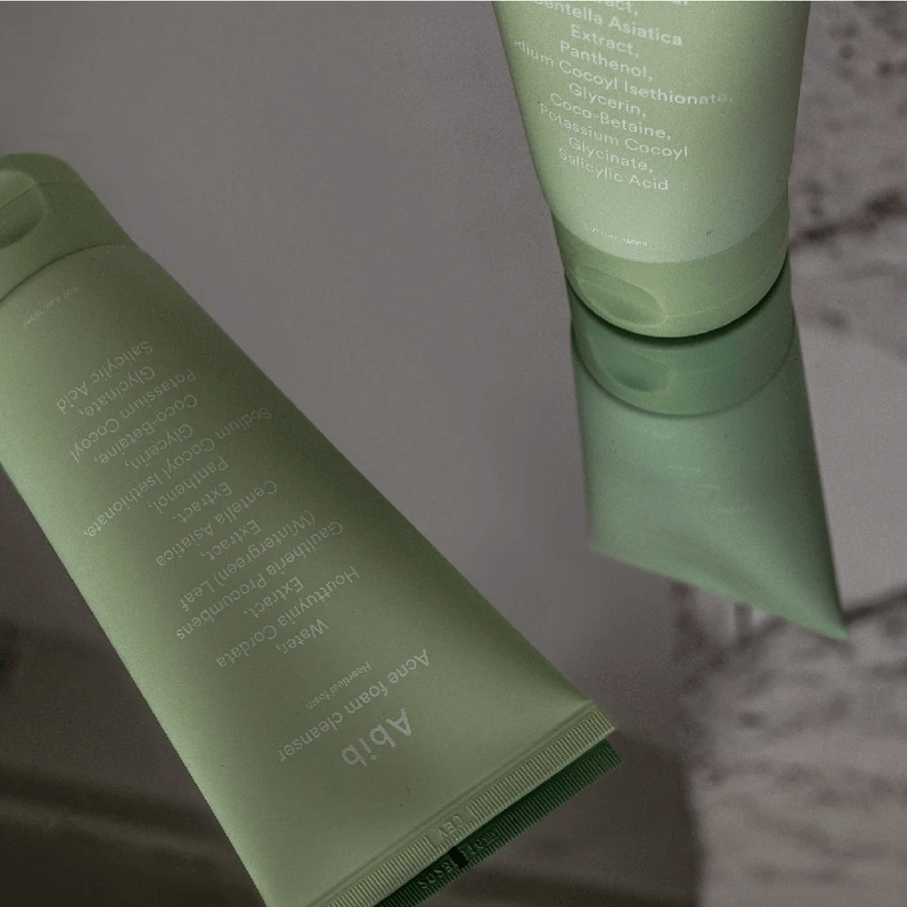 Tubo verde di Abib Acne Foam Cleanser Heartleaf sdraiato su un tavolo a specchio con la parte anteriore del prodotto e il testo riflessi. Gli ingredienti come l'acido salicilico, la centella e l'estratto di foglia di cuore sono chiaramente visibili sulla confezione.