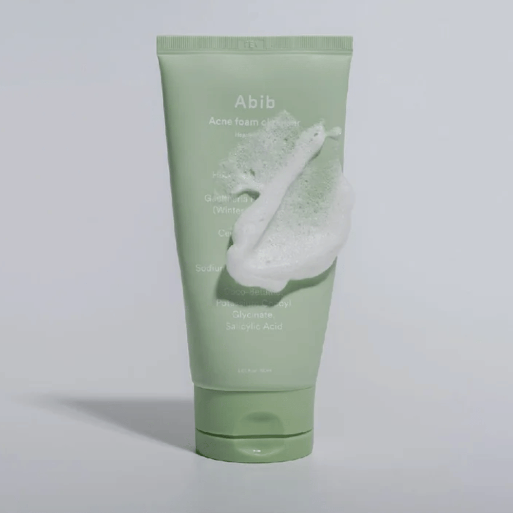 Tubo verde con Abib Acne Foam Cleanser Heartleaf, coperto da una schiuma bianca che illustra l'effetto detergente schiumoso. Il prodotto contiene estratto di foglia di cuore e acido salicilico per la pelle soggetta ad acne.