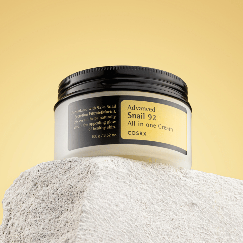 Un barattolo di COSRX Advanced Snail 92 All in one Cream posizionato su una superficie rocciosa con uno sfondo giallo.