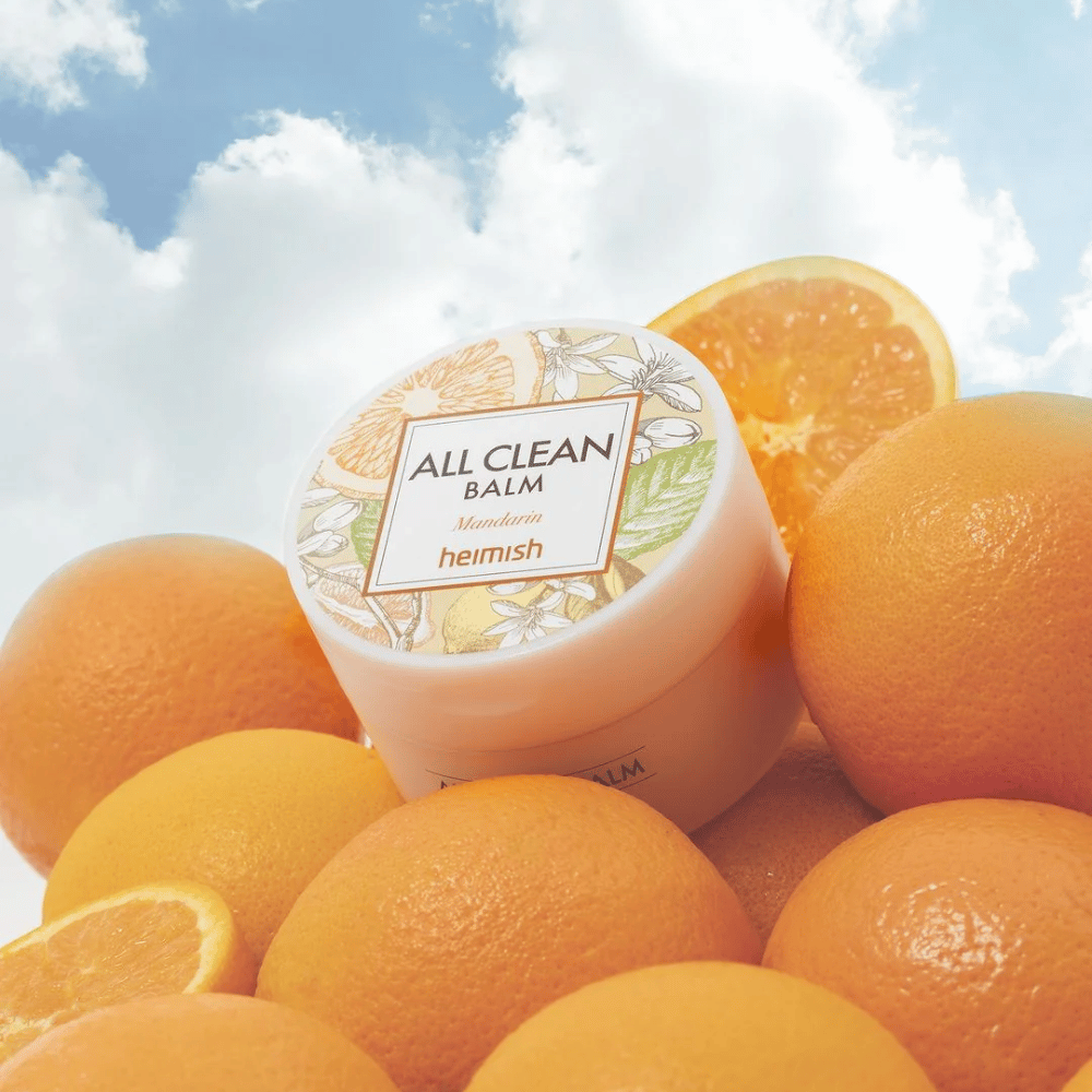 Barattolo di Heimish All Clean Balm Mandarin posizionato su un mucchio di arance, con un cielo blu sullo sfondo.