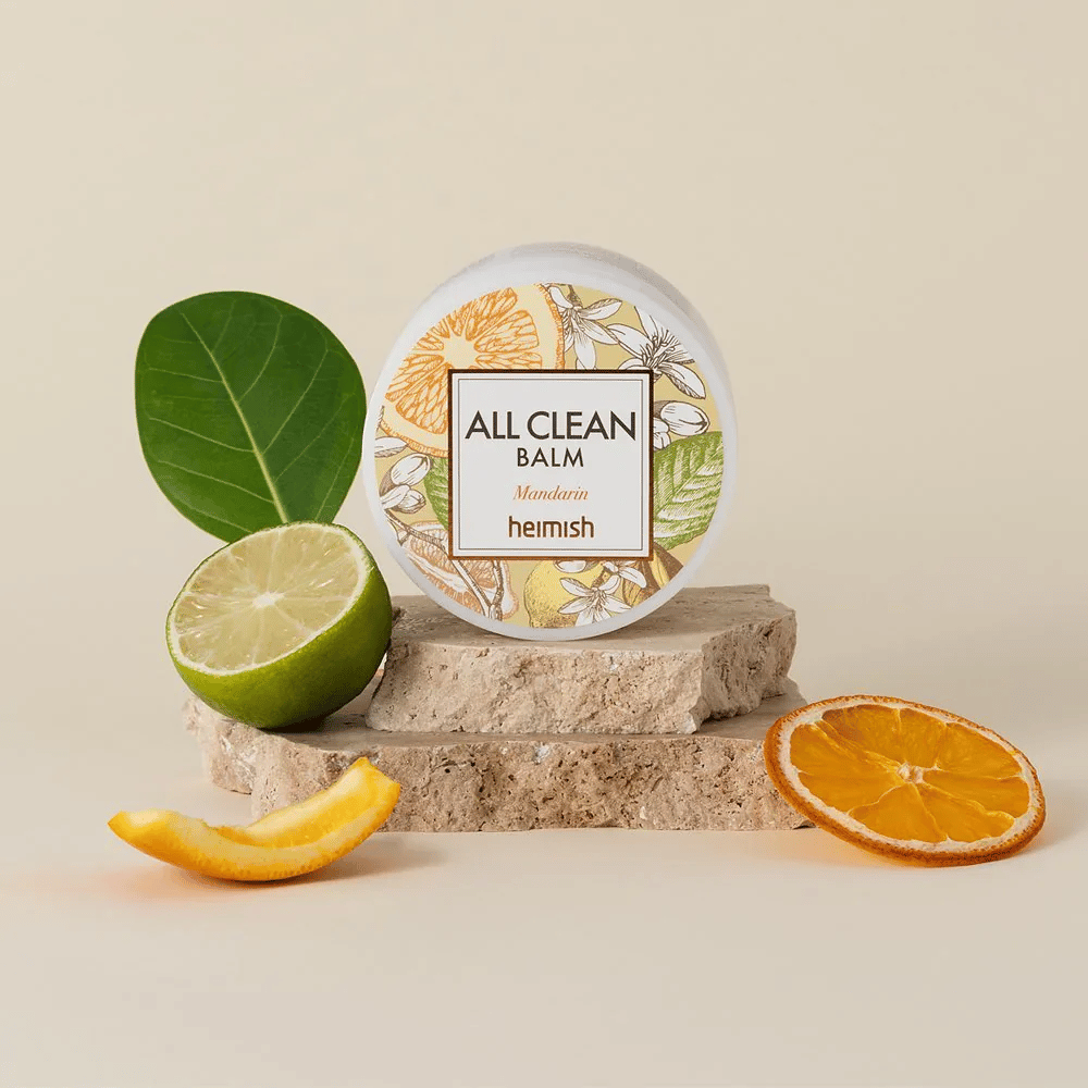 Barattolo di Heimish All Clean Balm Mandarin posizionato su pietre, circondato da bucce di mandarino e foglie di agrumi, davanti a uno sfondo chiaro.
