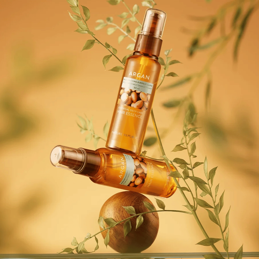 Due bottiglie Nature Republic Argan Essential Moisture Hair Oil posizionate tra foglie verdi – lucentezza setosa per capelli secchi.