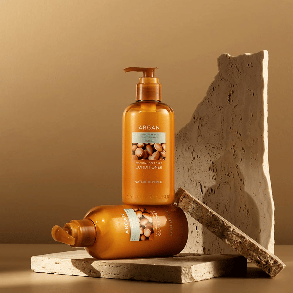 Due bottiglie di Nature Republic Argan Essential Moisture Repairing Conditioner su una superficie di pietra in tono caldo.