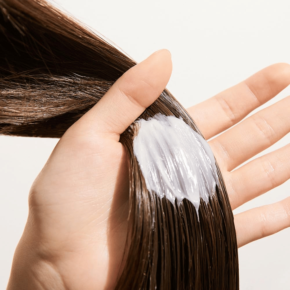 La mano applica Nature Republic Argan Essential Moisture Repairing Hair Mask su capelli bagnati – maschera idratante che rafforza e ammorbidisce i capelli.