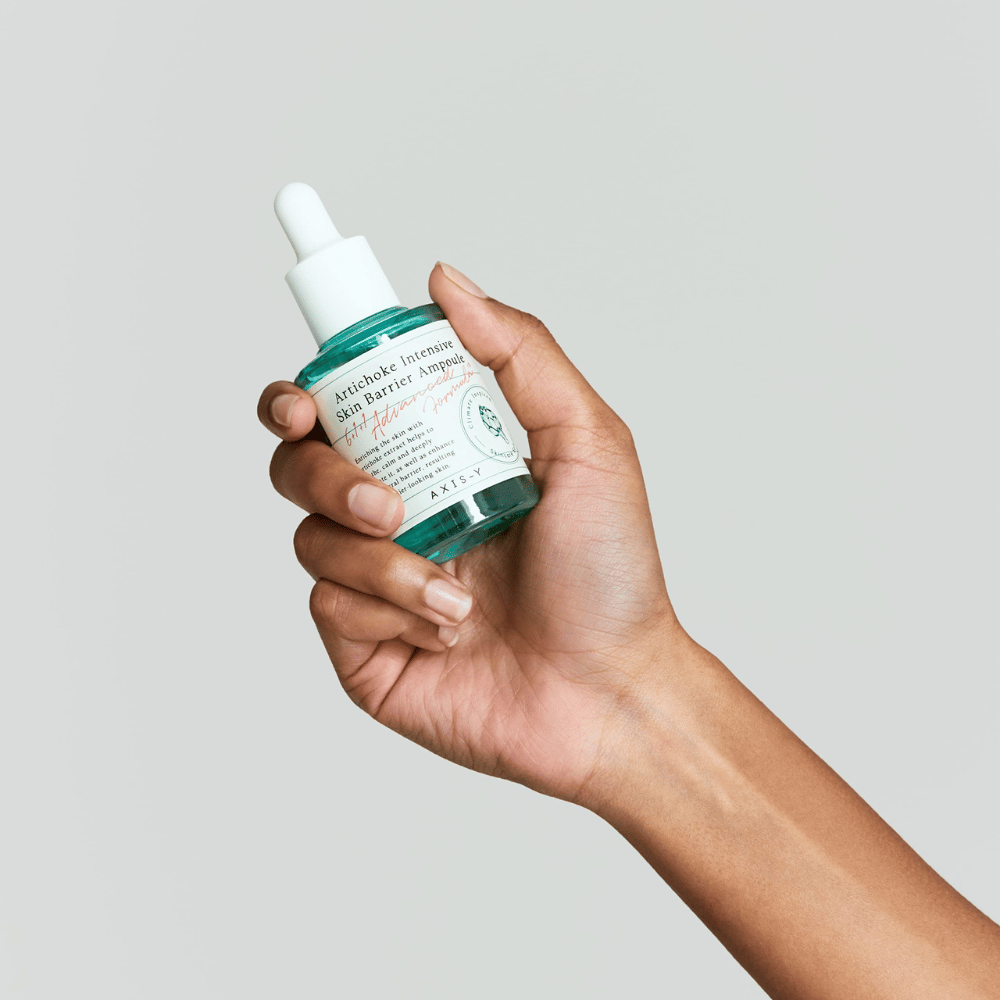 Una mano tiene una bottiglia di siero per la cura della pelle con l'etichetta "Artichoke Intensive Skin Barrier Ampoule.