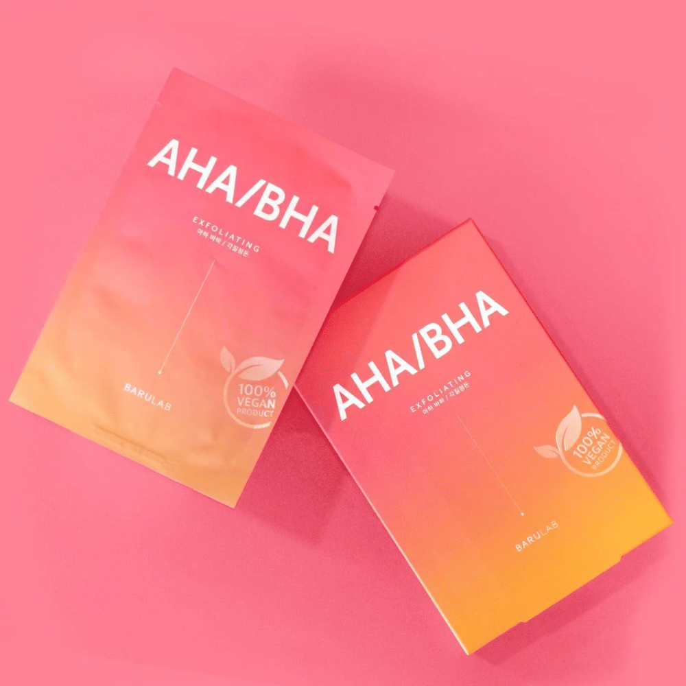 Due prodotti da BARULAB, uno in una busta e uno in una scatola, entrambi etichettati con "AHA/BHA" e "Exfoliating" su uno sfondo rosa. I prodotti sono contrassegnati come "100% Vegan Product".