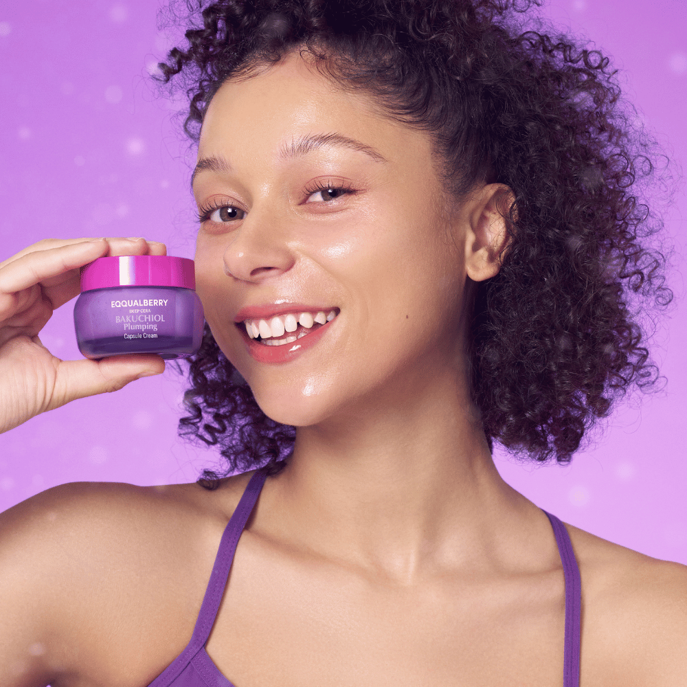 Donna con pelle luminosa tiene EQQUALBERRY Bakuchiol Plumping Capsule Cream e sorride davanti a uno sfondo viola.