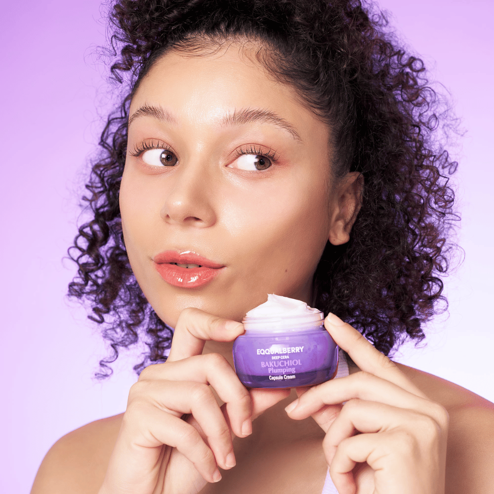 Donna tiene EQQUALBERRY Bakuchiol Plumping Capsule Cream in mano e mostra la sua pelle idratata e luminosa contro uno sfondo viola.