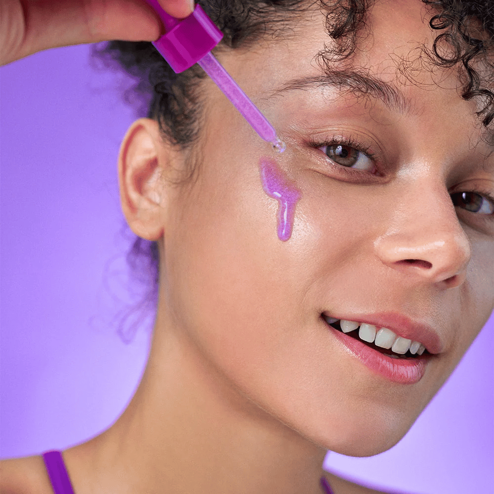 Donna sorridente applica un siero scintillante viola sulla pelle con una pipetta. Pelle radiosa e sfondo viola che si abbina alla tonalità del prodotto.
