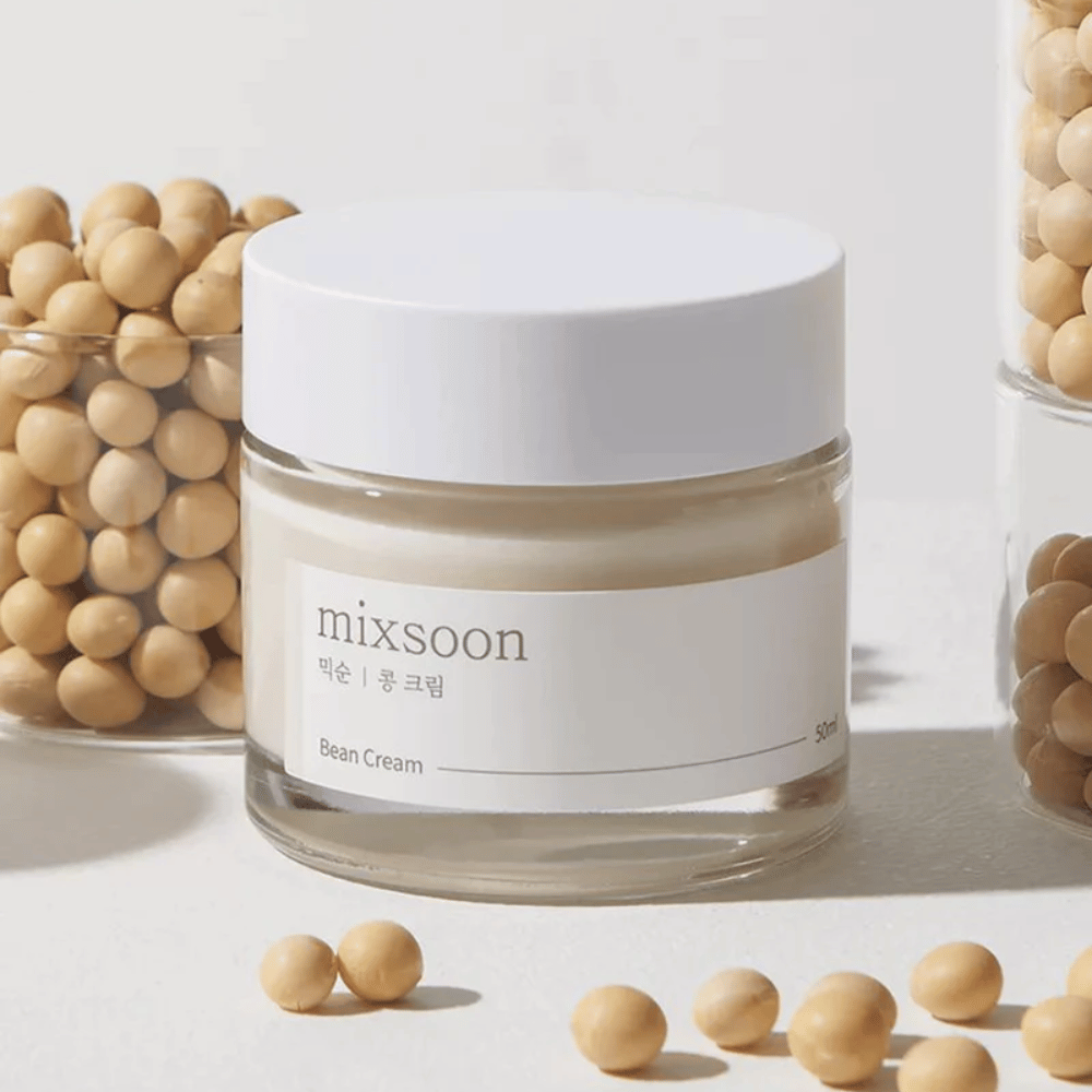 Un barattolo trasparente con etichetta e coperchio bianchi, etichettato "mixsoon 팥 크림" e "Bean Cream" con un volume indicato di "50ml". Il barattolo contiene una crema giallo chiaro ed è posizionato su una superficie chiara. Accanto al barattolo c'è un contenitore di vetro pieno di fagioli marrone chiaro rovesciati sulla superficie, suggerendo che la crema contiene estratto di fagioli. Lo sfondo e l'illuminazione creano un'atmosfera calda e naturale.