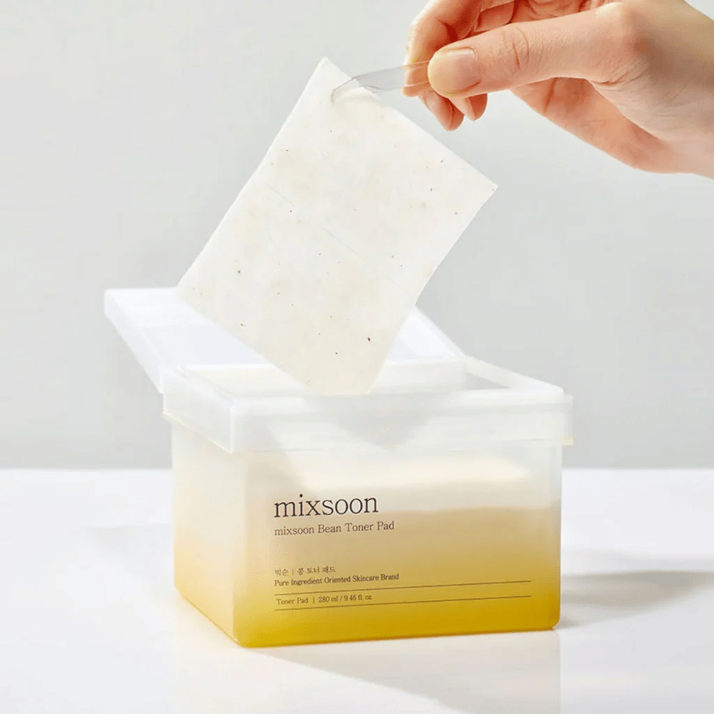 Una mano tiene una pinzetta e solleva un Mixsoon Bean Toner Pad da un contenitore quadrato e trasparente con un tono giallastro. Il contenitore contiene 70 dischetti tonici e ha testo che descrive il prodotto.