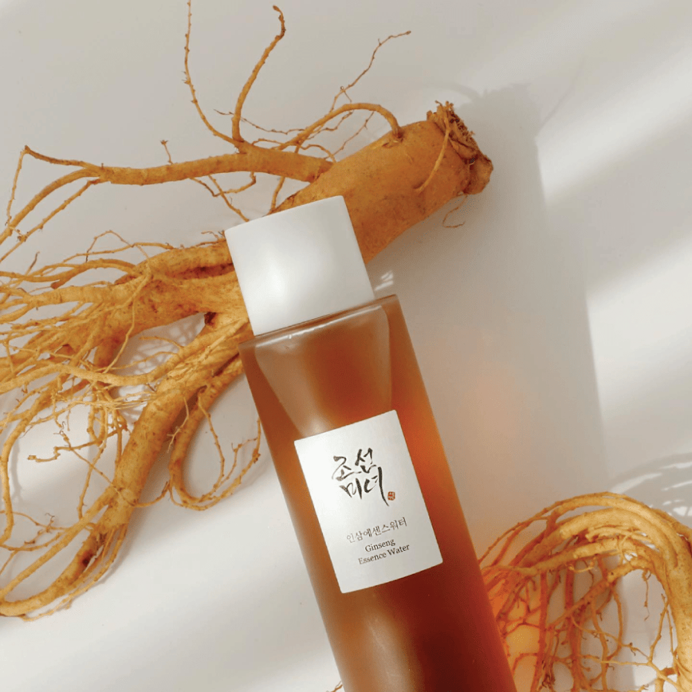 Una bottiglia marrone Beauty of Joseon Ginseng Essence Water con tappo bianco si trova accanto a una radice di ginseng fresca su sfondo bianco.