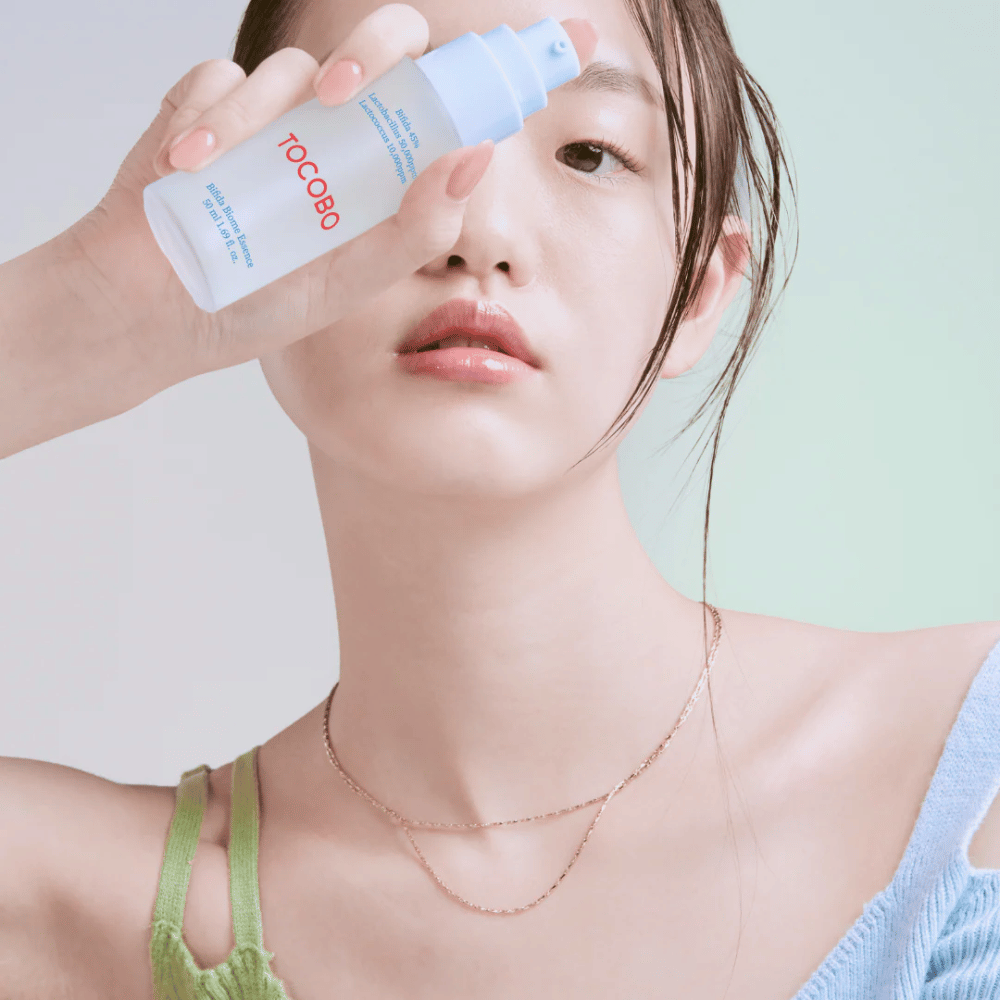 Giovane donna con trucco leggero e tono di pelle naturale tiene una bottiglia TOCOBO Bifida Biome Essence davanti al viso. Indossa una maglia azzurra con spalline verdi e una sottile collana d'oro su uno sfondo chiaro.