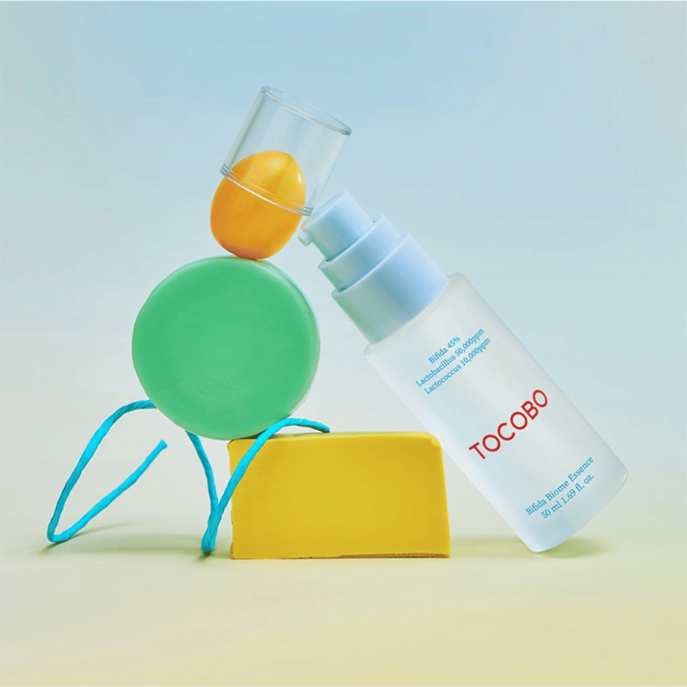 TOCOBO Bifida Biome Essence in un flacone a pompa smerigliato con logo rosso, posizionato su uno sfondo colorato con forme geometriche verdi, gialle e arancioni. Il testo sulla bottiglia indica Bifida 5%, Lactobacillus 3 000 ppm e Lactococcus 1 000 ppm.