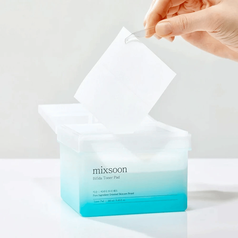 Un contenitore trasparente con una sfumatura blu e bianca che contiene Bifida dischetti tonici. Una mano prende un pad tonico viso dal contenitore, che è etichettato con "mixsoon Bifida Toner Pad" e contiene 280 ml (9,46 fl. oz.) di dischetti tonici destinati alla cura della pelle.