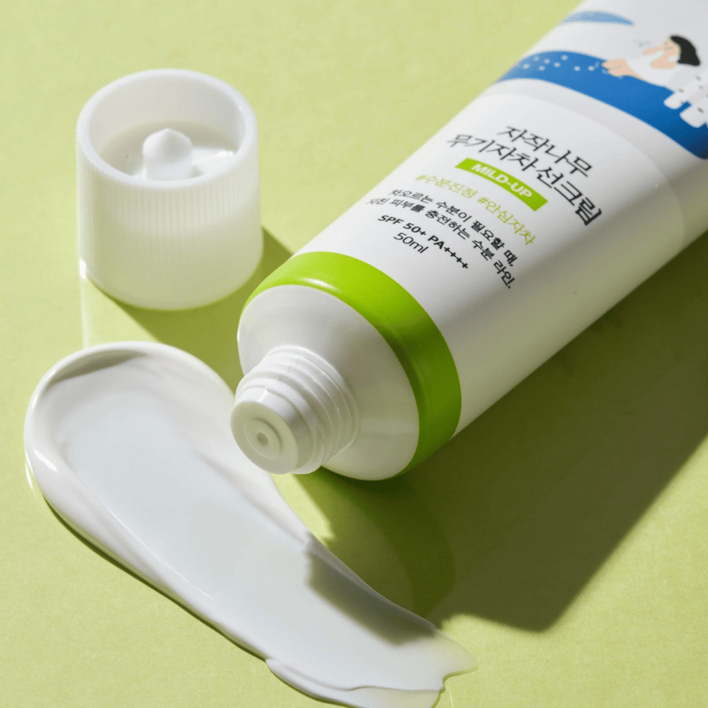 Primo piano di Birch Juice Moisturizing Mild Up Sunscreen con tappo svitato, crema solare bianca spalmata su sfondo verde chiaro, SPF 50+ PA++++, 50 ml.