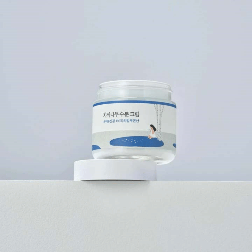 Round Lab Birch Juice Moisturizing Cream in barattolo trasparente con etichetta bianca e illustrazione blu di donna presso betulla, posizionato su superficie bianca contro sfondo chiaro.