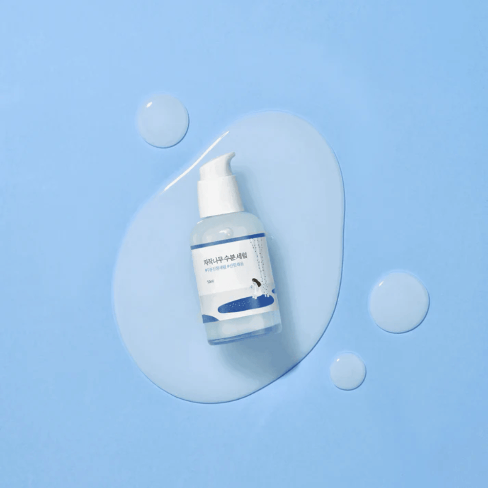 Round Lab Birch Juice Moisturizing Serum in flacone con pompa trasparente posizionato su uno sfondo blu con gocce e siero versato.