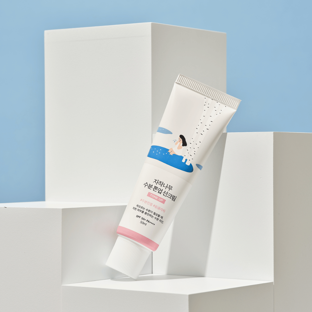 Un tubo con Birch Juice Moisturizing Tone Up Sunscreen posizionato su cubi bianchi contro uno sfondo azzurro. Il tubo è bianco con un'illustrazione di una donna vicino a una betulla e acqua. Il testo in coreano indica che il prodotto ha SPF 50+ PA++++ e contiene 50 ml.
