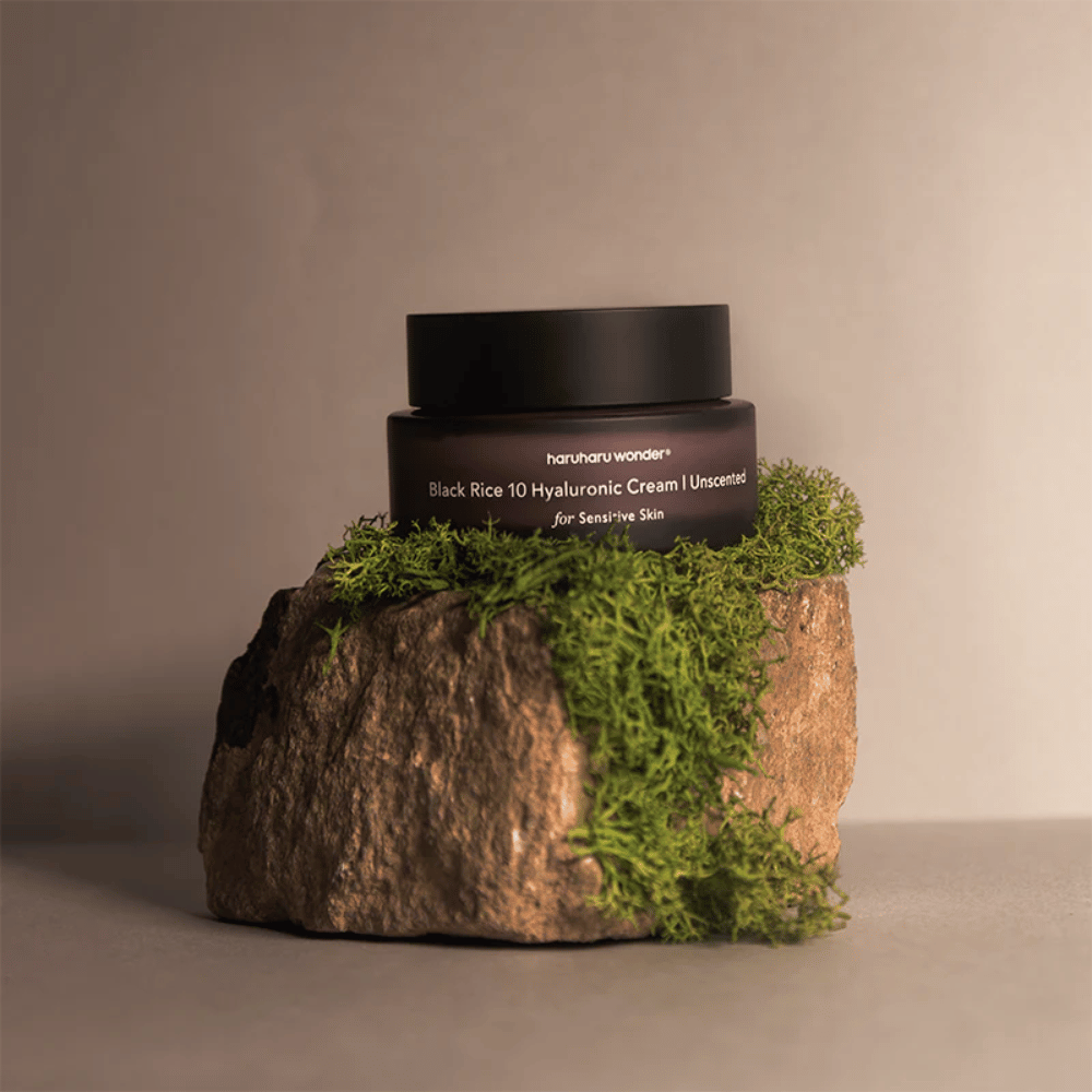 L'immagine mostra un barattolo di crema per la pelle etichettato "haruharu wonder Black Rice 10 Hyaluronic Cream Unscented for Sensitive Skin". Il barattolo è nero e si trova su una grande pietra coperta di muschio verde chiaro, creando una sensazione naturale e terrosa.