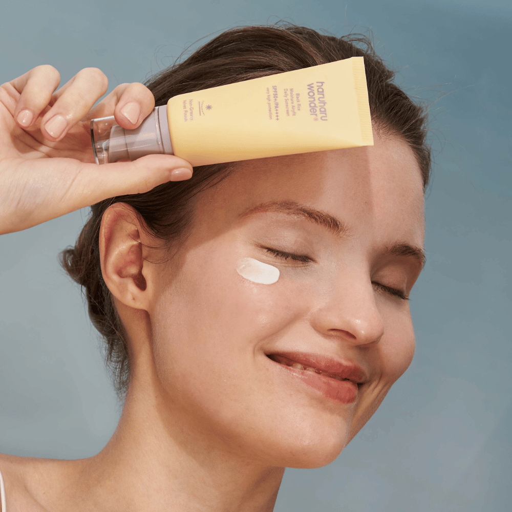 Una donna sorridente applica Black Rice Moisture Airyfit Daily Sunscreen SPF 50+ PA++++ da Haruharu Wonder sul suo viso. Tiene in mano un tubo giallo con crema solare e ha un po' di prodotto sulla guancia. Lo sfondo è azzurro chiaro.