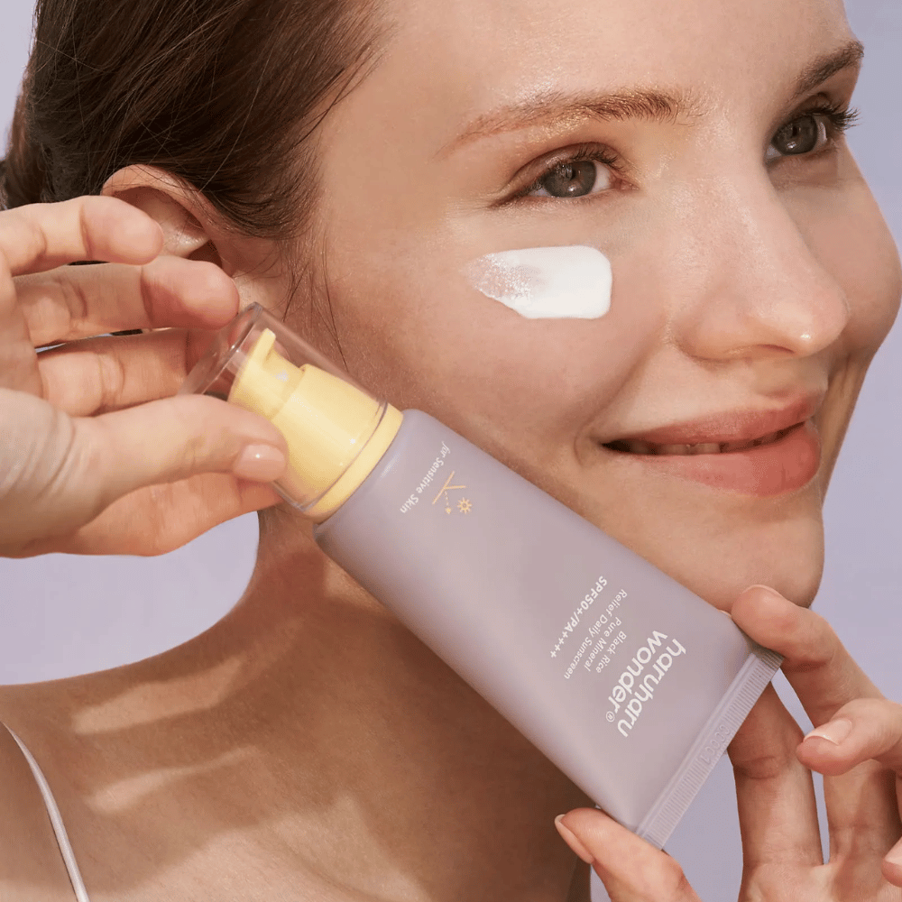 Una donna sorridente applica Haruharu Wonder Black Rice Pure Mineral Relief Daily Sunscreen SPF 50+ PA++++ sul viso. Tiene un tubo viola con crema solare e ha una goccia del prodotto sulla guancia. Lo sfondo è lilla chiaro.