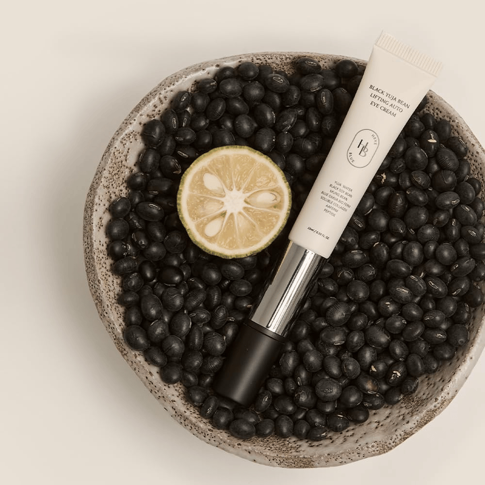 Black Yuja Bean Lifting Auto Eye Cream adagiato in una ciotola piena di fagioli neri e un agrume tagliato a metà - una rappresentazione visiva degli ingredienti naturali del prodotto.