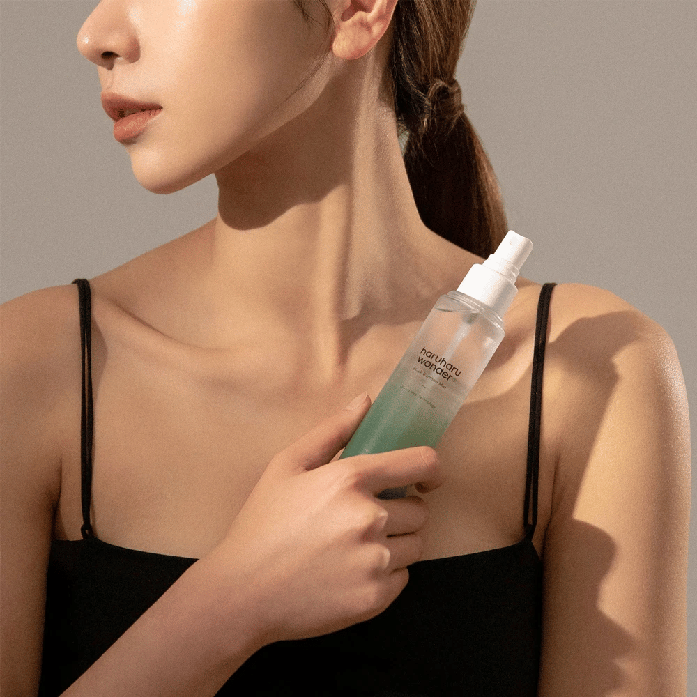 Immagine di HaruHaru Wonder Black Bamboo Mist tenuta da una donna. Il prodotto è in una bottiglia spray trasparente con una tonalità verde e una pompa bianca. La donna ha un aspetto rilassato, indossa un top nero ed è concentrata sull'utilizzo della nebbia per dare idratazione e luminosità alla pelle. La bottiglia fa parte di una routine di cura della pelle.