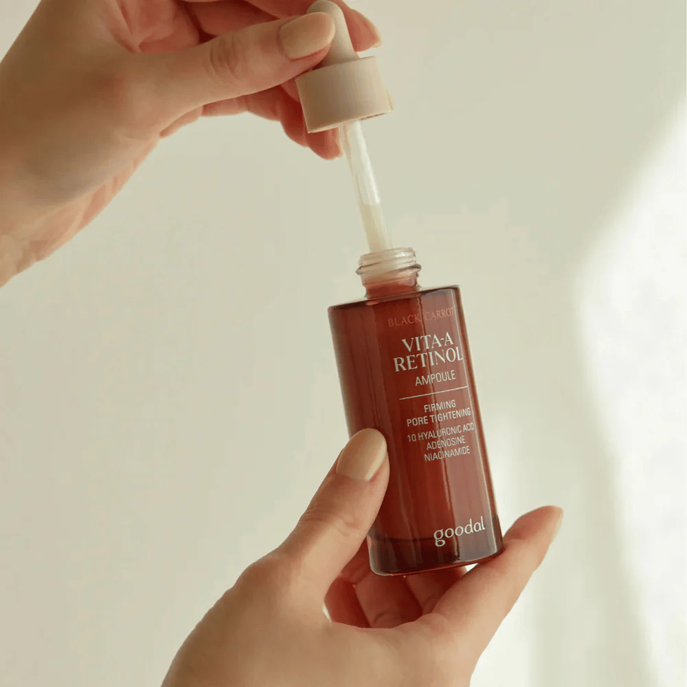 Una mano tiene una bottiglia con Goodal Black Carrot Vita-A Retinol Firming Ampoule. L'etichetta della bottiglia mostra il nome del prodotto e le caratteristiche, inclusi "Firming Pore Tightening" e ingredienti come 10% di acido ialuronico, adenosina e niacinamide. Il contagocce è di colore beige.