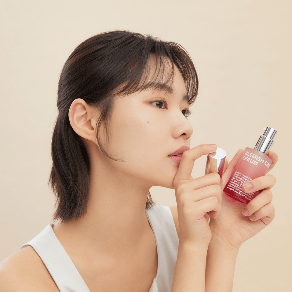 Una donna con capelli scuri tiene una bottiglia di Blemish Care Serum in una mano e rimuove il tappo con l'altra. Ha un'espressione facciale neutra e lo sfondo è beige chiaro, dando un'impressione morbida e minimalista.