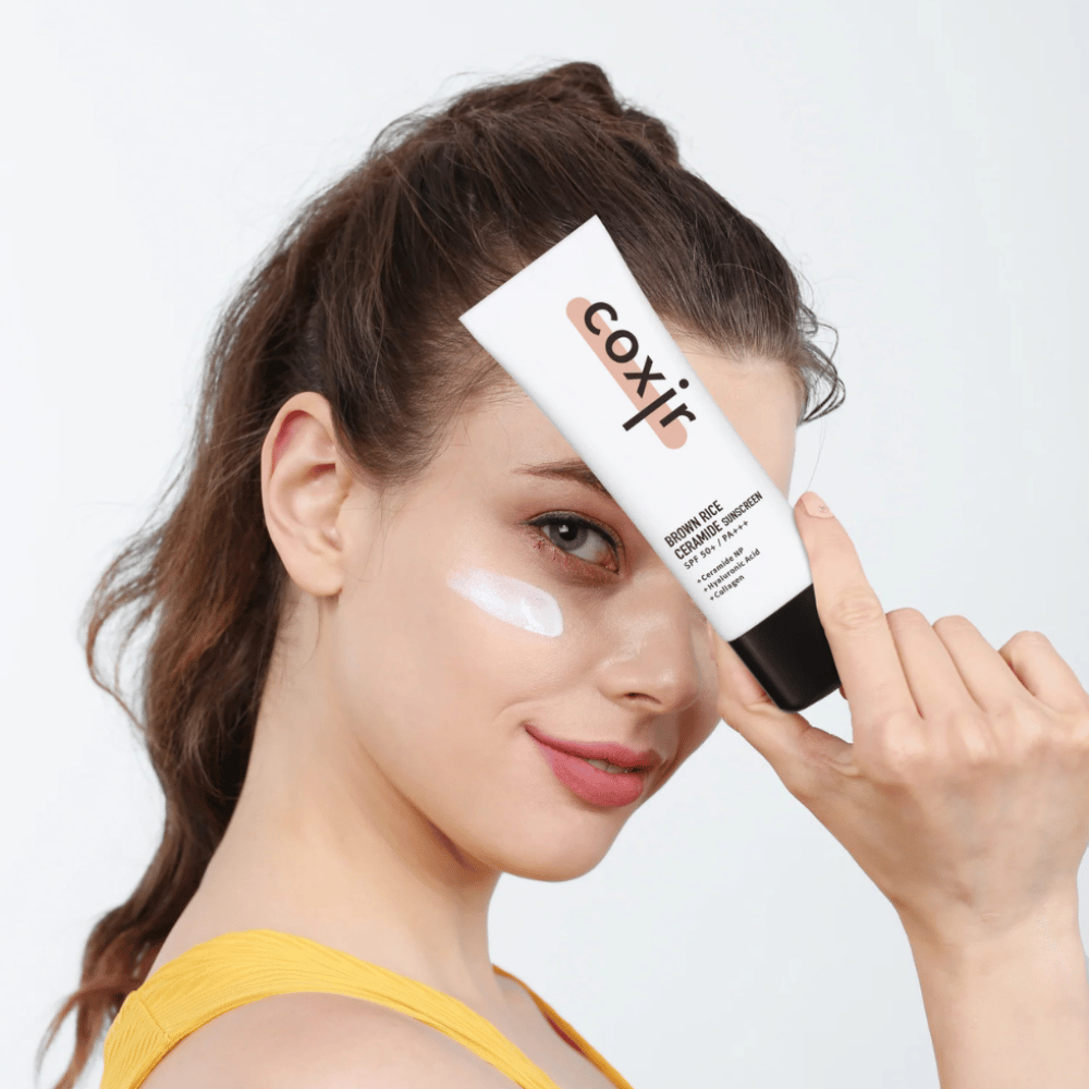 Donna con crema solare applicata sulla guancia tiene un tubo Coxir Brown Rice Ceramide Sunscreen, SPF 50+ PA+++, contro la fronte e sorride leggermente.