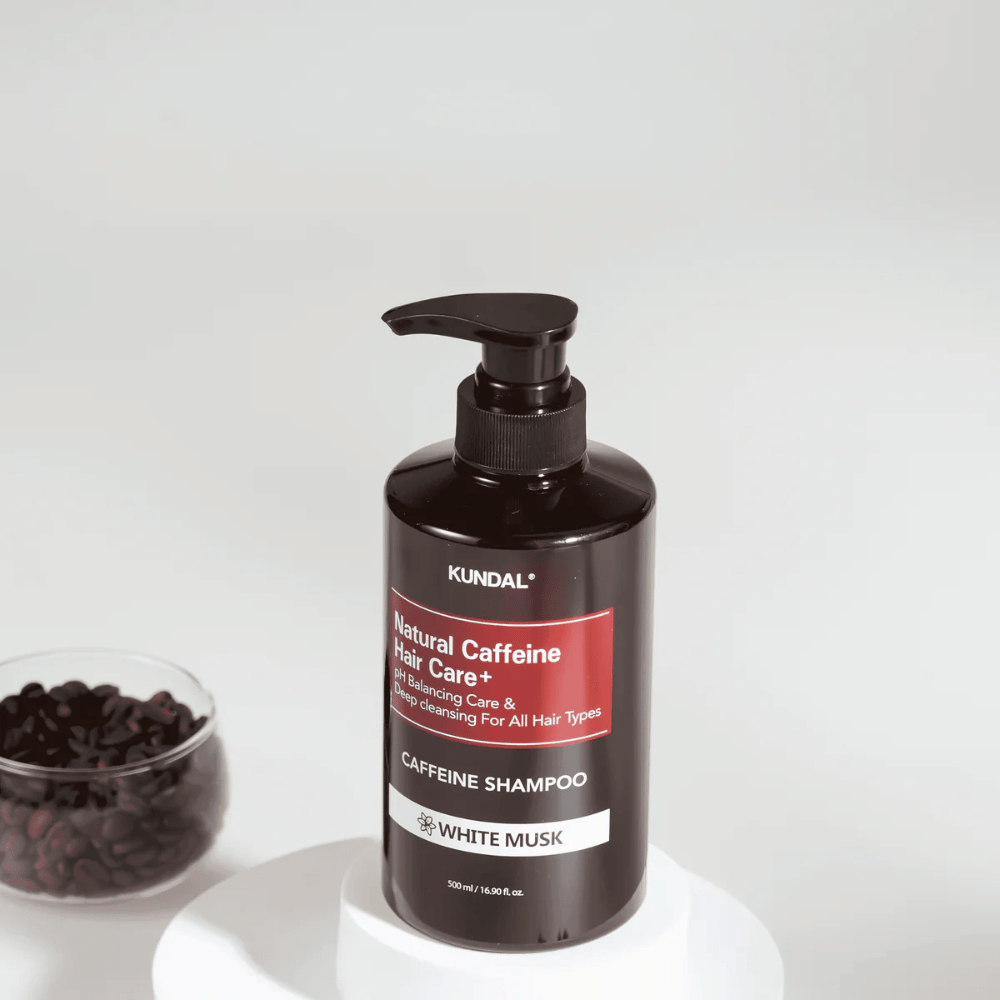 KUNDAL Natural Caffeine Hair Care+ Shampoo White Musk in bottiglia con pompa nera, 500 ml, posizionata accanto a una ciotola di chicchi di caffè.