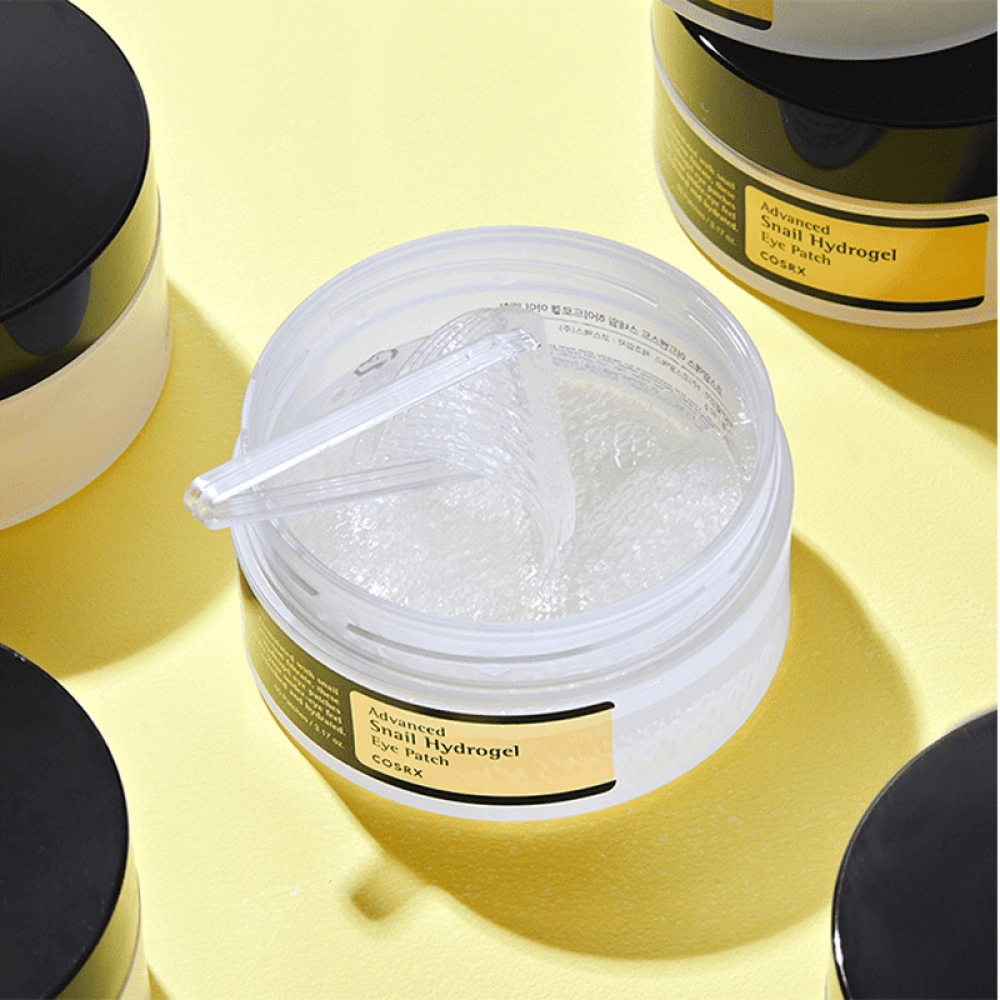 Un barattolo aperto COSRX Advanced Snail Hydrogel Eye Patch con patch per occhi trasparenti e una piccola spatola, circondato da altri barattoli chiusi su sfondo giallo.