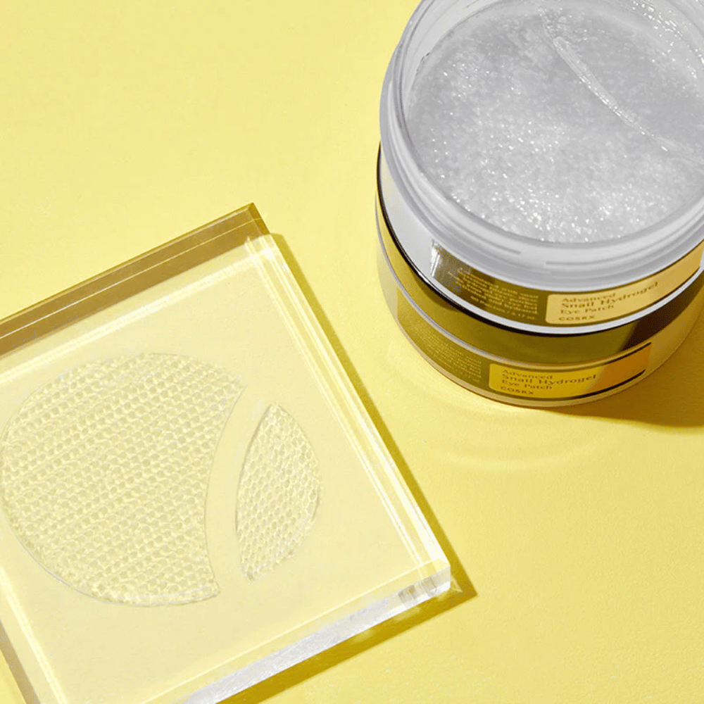 Un barattolo aperto COSRX Advanced Snail Hydrogel Eye Patch si trova su uno sfondo giallo accanto a un sottobicchiere trasparente dove sono posizionate due patch per occhi.