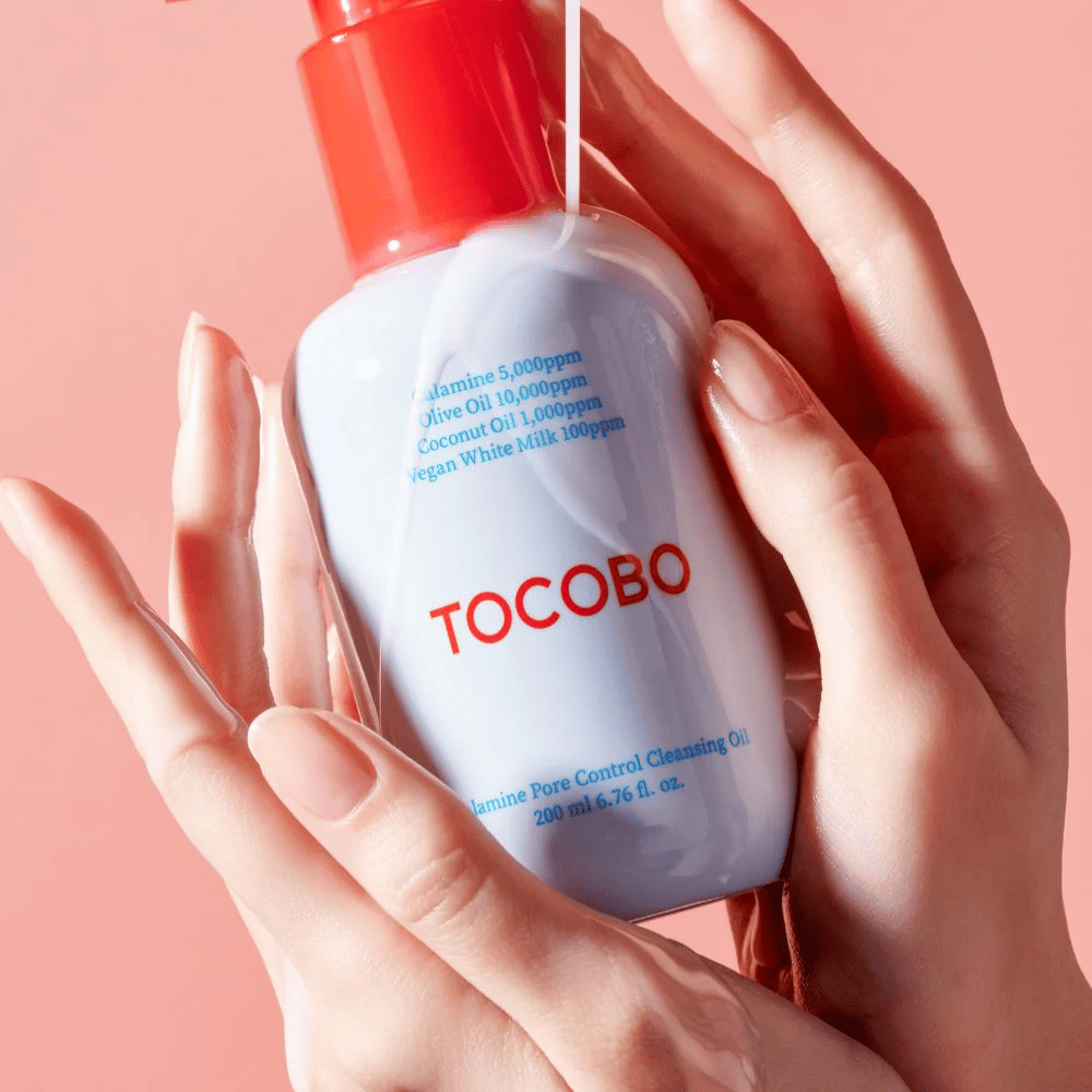 TOCOBO Calamine Pore Control Cleansing Oil tenuto in mano su uno sfondo rosa, il dosatore rosso del flacone e la lista degli ingredienti sono chiaramente visibili.
