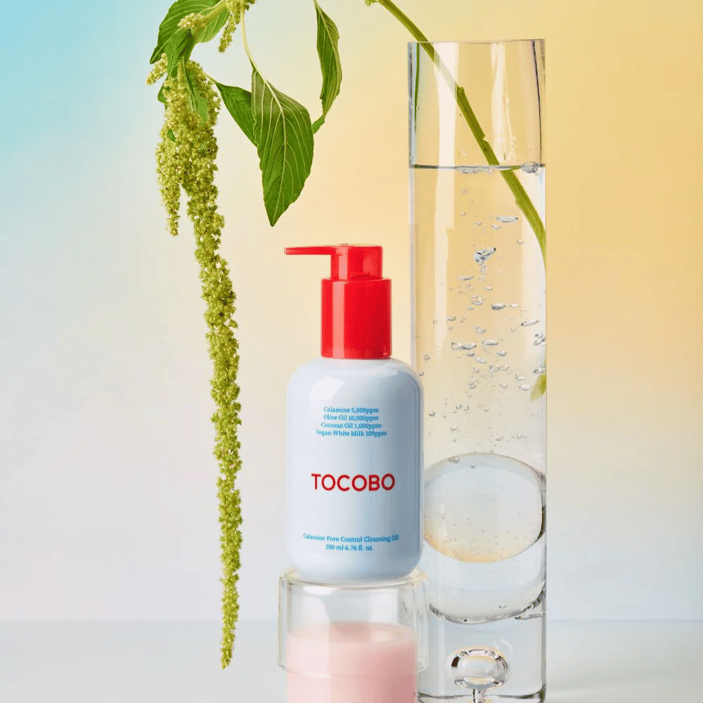 TOCOBO Calamine Pore Control Cleansing Oil con dosatore rosso si trova accanto a un alto tubo di vetro riempito d'acqua e una pianta verde, su uno sfondo dai colori pastello chiari.