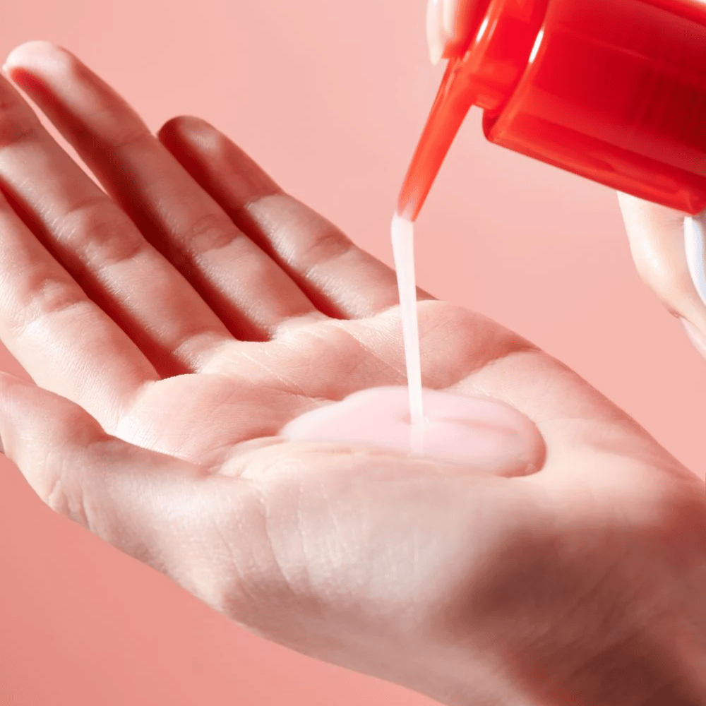 Un olio detergente rosa chiaro viene applicato da un flacone dosatore rosso in una mano aperta su uno sfondo rosa.