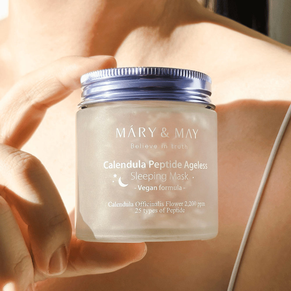 Una donna tiene un barattolo di MARY & MAY Calendula Peptide Ageless Sleeping Mask. Il barattolo è trasparente con un coperchio viola e il testo "Calendula Peptide Ageless Sleeping Mask" è chiaramente visibile. Il prodotto è una maschera per dormire vegana con calendula e peptidi, progettata per dare alla pelle una luminosità giovanile durante la notte.