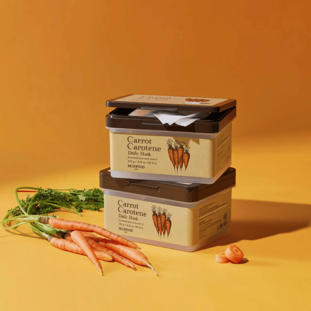 L'immagine mostra tre confezioni impilate di "Carrot Carotene Daily Mask" da SKINFOOD, posizionate su uno sfondo arancione insieme a carote fresche. Ogni scatola contiene maschere facciali arricchite con β-carotene dall'olio di semi di carota, progettate per l'uso quotidiano per nutrire e curare la pelle.